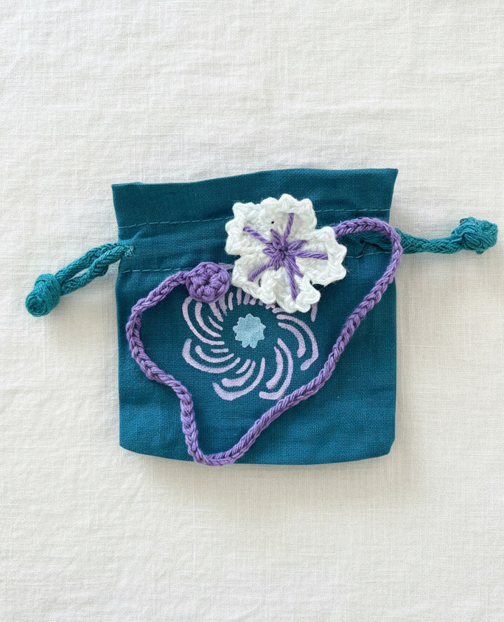 Gehäkelte Blume mit violetter Kordel auf blauem Stoff-Säckchen mit Spiralmotiv auf leinentuch Hintergrund