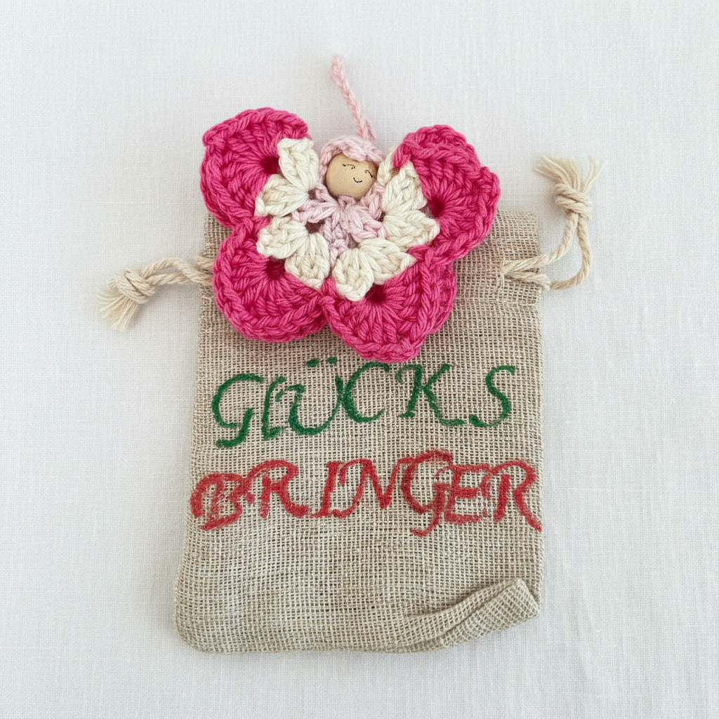 Handgehäkelter Schmetterling Glücksbringer in Rosa und Creme auf kleinem Stoffbeutel mit der Aufschrift „Glücksbringer“ – liebevoll handgemacht von Regenbogen Creations 💛