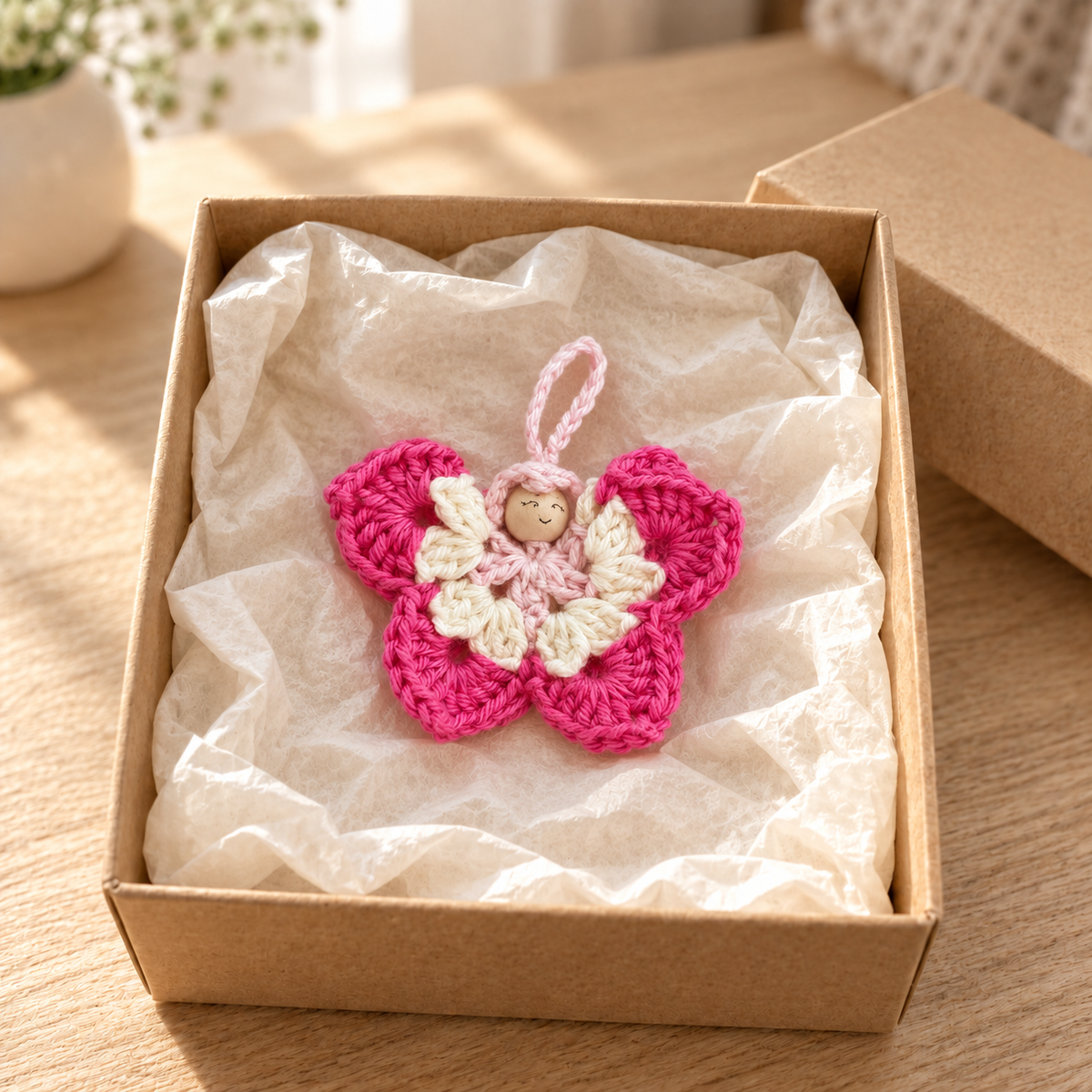 Kleiner gehäkelter Schmetterling in Pink und Creme mit Holzperlenkopf auf weissem Seidenpapier in einer offenen Geschenkschachtel.