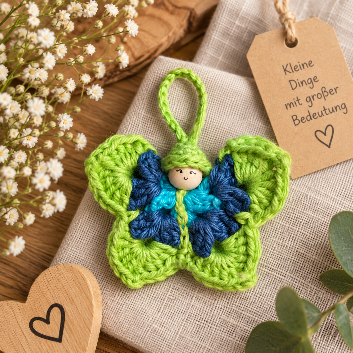 Handgemachter gehäkelter Schmetterling in Grün, Blau und Türkis mit Holzkopf und Aufhängeschlaufe, liebevoll auf Leinen dekoriert.