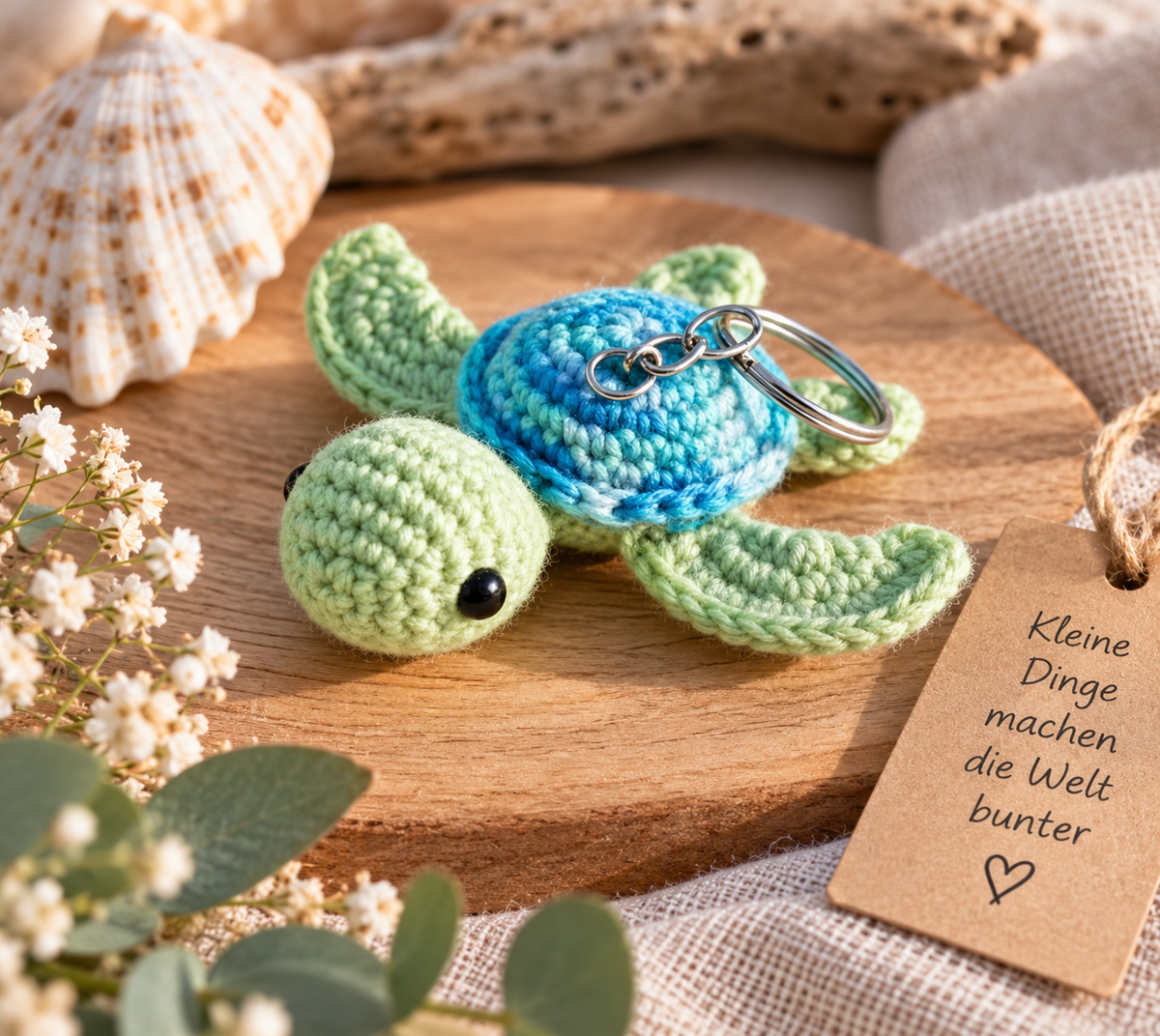 Handgehäkelte Schildkröte als Schlüsselanhänger in Grün und Türkis, dekoriert mit Muschel, Blumen und Holzanhänger.
