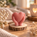 Handgemachte rosa Herzkerze in schützenden Händen auf Holzscheibe in gemütlicher Dekoration.