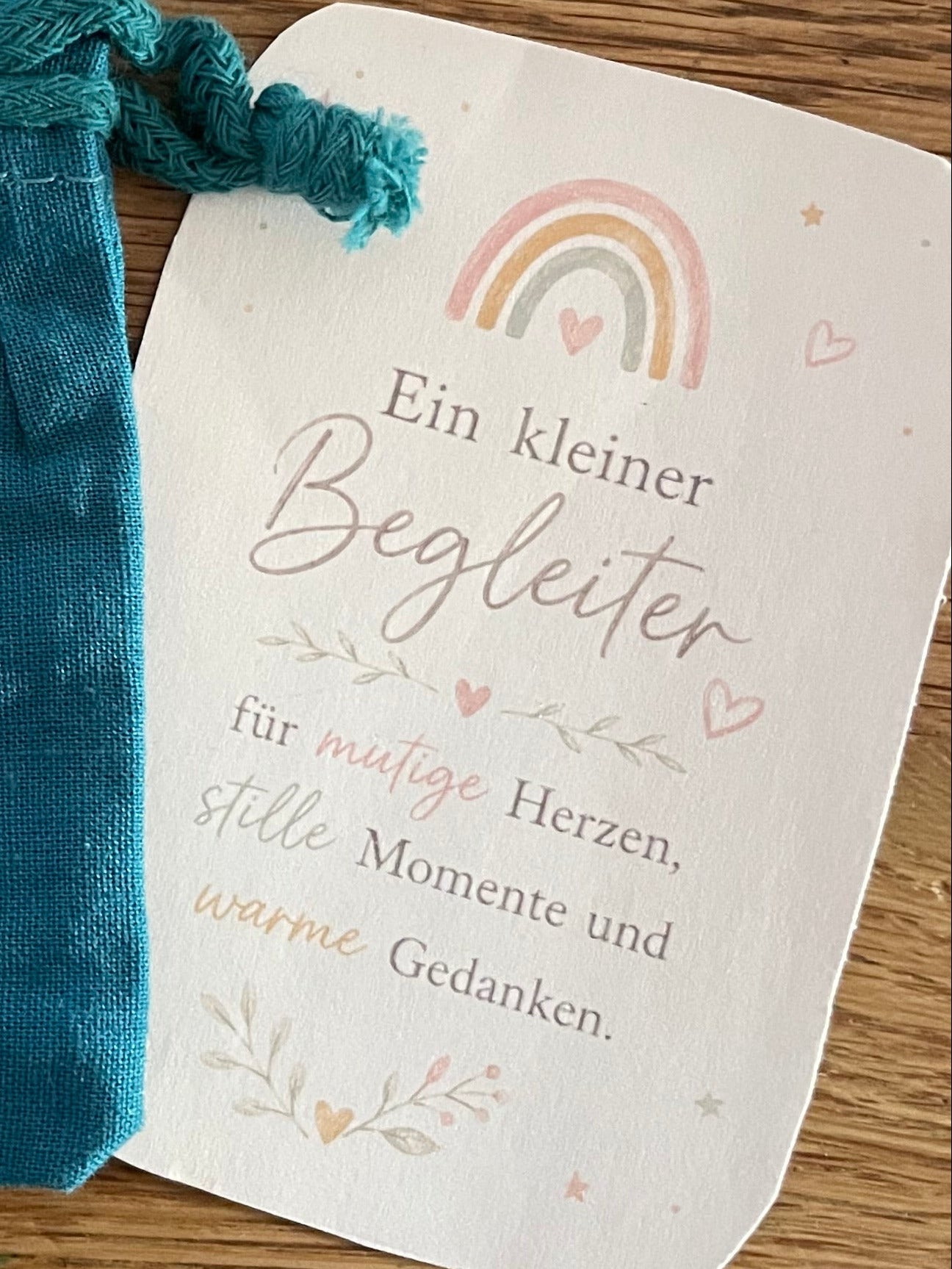 Kärtchen mit Regenbogen und Aufschrift „Ein kleiner Begleiter“ neben blauem Stoff-Säckchen auf Holztisch.