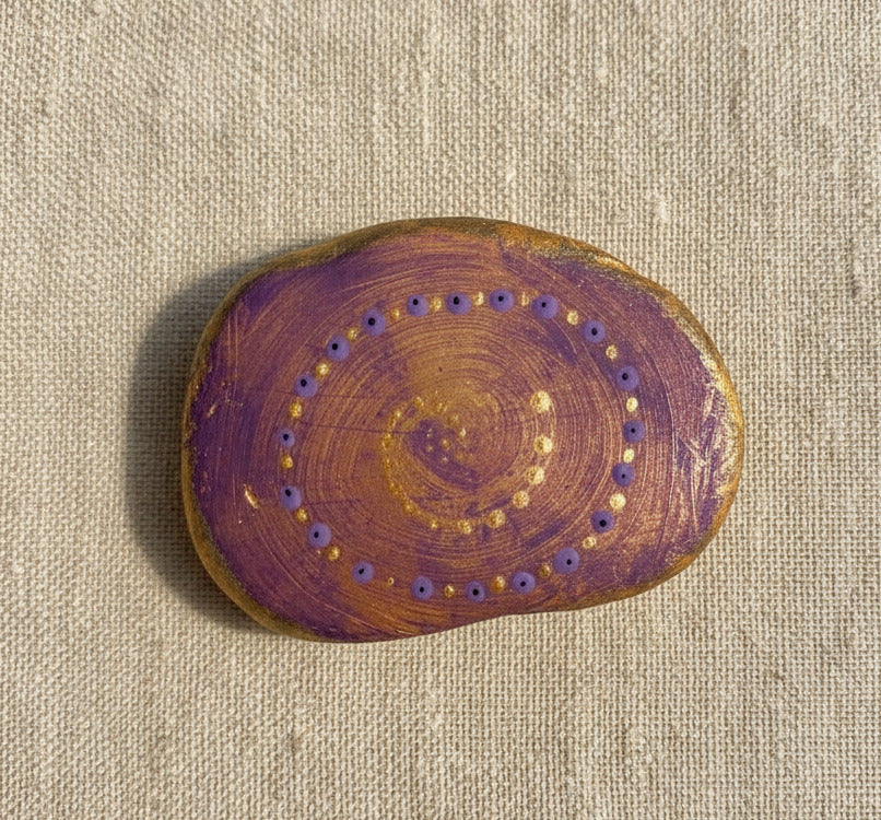 Handbemalter Mandala-Spirale-Stein in Lila und Gold mit feinen Punktmustern auf hellem Hintergrund