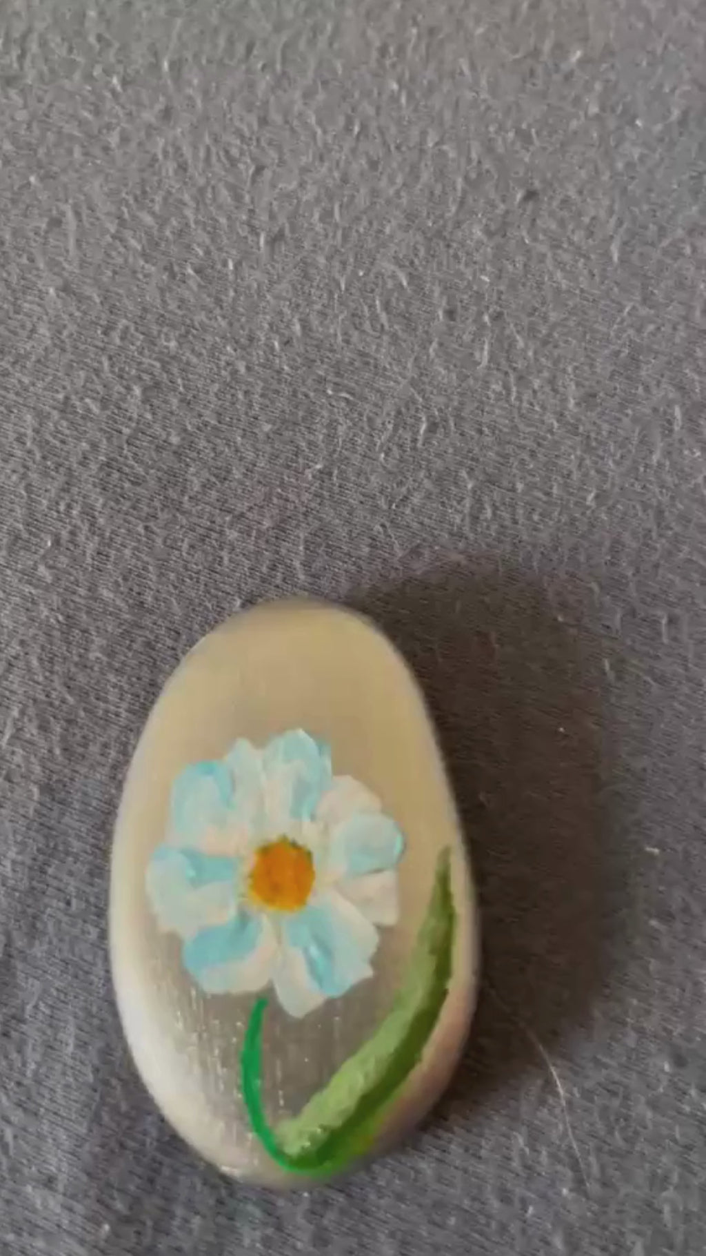 Hand bemalter Glücksstein mit hellblauer Blume und grünen Blättern. Der Stein wird in der Hand gehalten und in einem kurzen Video von verschiedenen Seiten gezeigt.