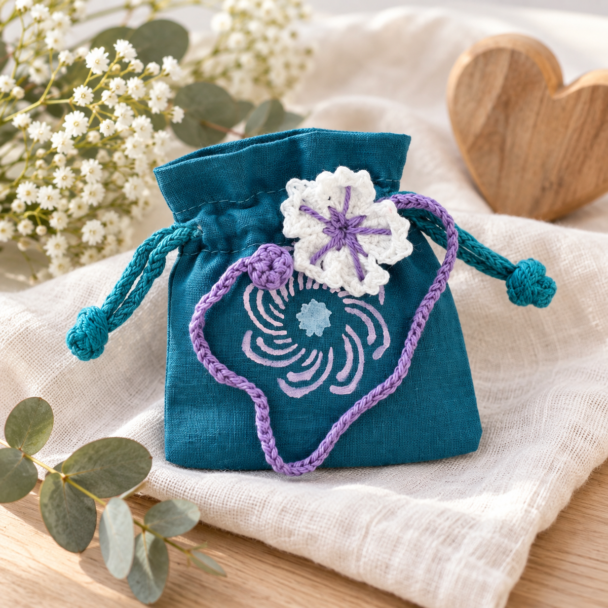 Türkises Stoffbeutelchen mit gehäkeltem Blumenanhänger in Lila und Weiß, dekoriert mit Blumen und Eukalyptus.